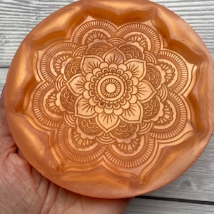May include: A round, orange-toned resin coaster with a detailed mandala design. The coaster features a raised floral pattern in a circular shape, with a slightly raised edge. The color is a warm, metallic orange.