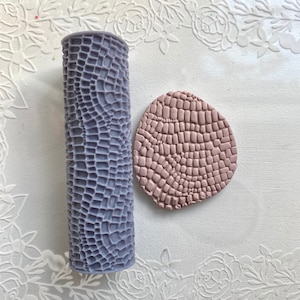 Puede incluir: Un rodillo de silicona morado con un patrón texturizado de piel de cocodrilo y una impresión de arcilla rosa del mismo patrón.