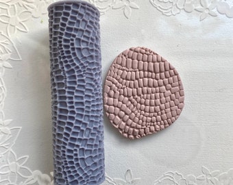 May include: A purple silicone rolling pin with a textured alligator skin pattern and a pink clay impression of the same pattern.