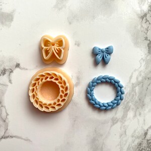 May include: Four cookie cutters: a wreath in a light brown colour, a wreath in a light blue colour, a butterfly in a light brown colour, and a bow in a light blue colour.