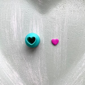 May include: A teal heart-shaped cookie cutter and a small pink heart on a light grey background.