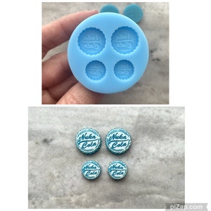 May include: A silicone mould with four impressions of a Nuka Cola bottle top. There are four finished bottle tops in blue and white with the text "Nuka Cola" on them.