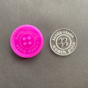 Puede incluir: Un molde de silicona rosa y un cortador de galletas de acrílico transparente, ambos con forma de botón con el texto "Polo Norte" y "Santa Claus" alrededor del botón.