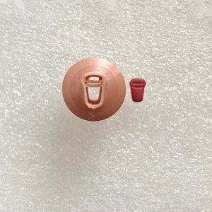 May include: A close-up of a copper-colored cookie cutter shaped like a coffee cup. A smaller, red-colored coffee cup is shown to the right of the cutter.