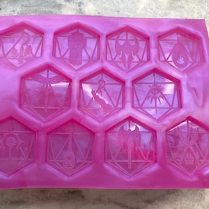May include: A pink silicone mold with 10 hexagonal cavities, each featuring a different Dungeons and Dragons dice symbol. The symbols include a dragon, a fist, a sword, a hand, a sun, a lute, an axe, and a pair of crossed swords.