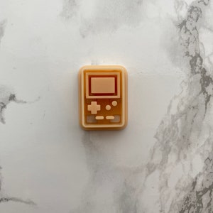 May include: A beige plastic cookie cutter shaped like a handheld video game console. The cookie cutter has a screen, buttons, and a directional pad.