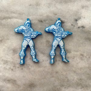 May include: Two blue plastic charms featuring a white outline of a cartoon character holding a sword. The character is wearing a helmet and a loincloth.