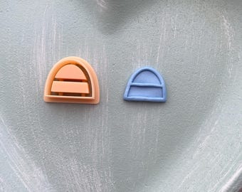 Puede incluir: Dos cortadores de galletas arqueados, uno naranja y otro azul, se encuentran sobre una superficie en forma de corazón verde claro. El cortador naranja tiene dos líneas horizontales, el azul una.