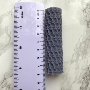 Könnte beinhalten: Eine graue, strukturierte Rolle mit einer Länge von etwa 6 cm. Die Rolle hat ein sich wiederholendes Muster aus kleinen, erhabenen Punkten.