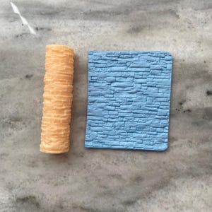 May include: Two silicone cake decorating tools. One is a blue rectangular sheet with a raised stone pattern. The other is a beige cylinder with a raised brick pattern.