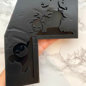 May include: Black acrylic Halloween decoration with a witch, pumpkin, and ghost cutouts. The witch wears a pointed hat and holds a pumpkin. The ghost has a smiling face. The decoration is L-shaped.