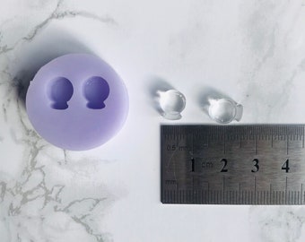 May include: A purple silicone mould with two round cavities for making small round objects. Two clear plastic round objects are next to a ruler measuring 1 cm.