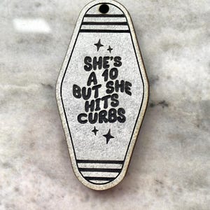 May include: White motel key tag with black text that reads "She's a 10 but she hits curbs".