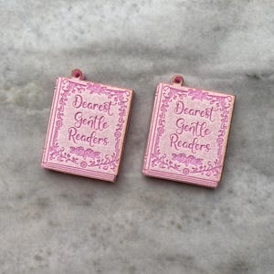 Puede incluir: Dos colgantes rosas con forma de libro con el texto "Dearest Gentle Readers". Los colgantes tienen un borde floral y un peque&ntilde;o lazo en la parte superior. Ideales para la creaci&oacute;n de joyas.