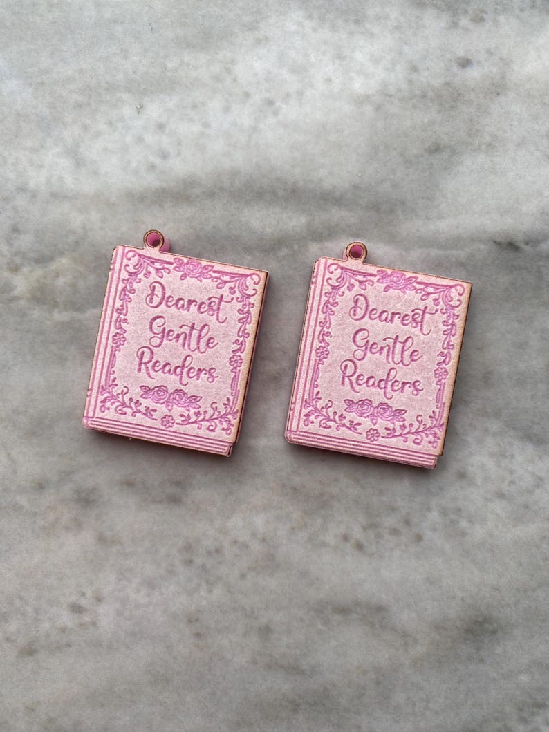 Puede incluir: Dos colgantes rosas con forma de libro con el texto "Dearest Gentle Readers". Los colgantes tienen un borde floral y un peque&ntilde;o lazo en la parte superior. Ideales para la creaci&oacute;n de joyas.