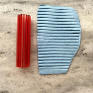May include: A red plastic tool with a textured surface and a blue plastic tool with a textured surface. Both tools are used for creating patterns in clay.