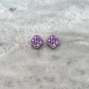 May include: Two purple clover-shaped stud earrings with a geometric design. The earrings have a light purple border and a darker purple interior, set against a marble background. The earrings are small and delicate.