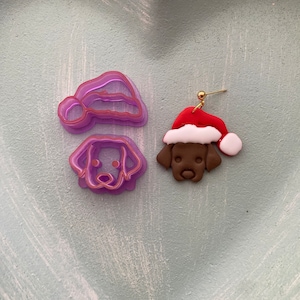 May include: A pair of Christmas-themed earrings featuring a brown dog wearing a red and white Santa hat. The earrings are made of polymer clay and have a gold hook.