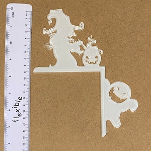 May include: Clear acrylic Halloween corner decoration. The design includes a witch with a pointed hat, a jack-o'-lantern, and a ghost. The decoration is designed to fit around a corner and is approximately 20 cm long.