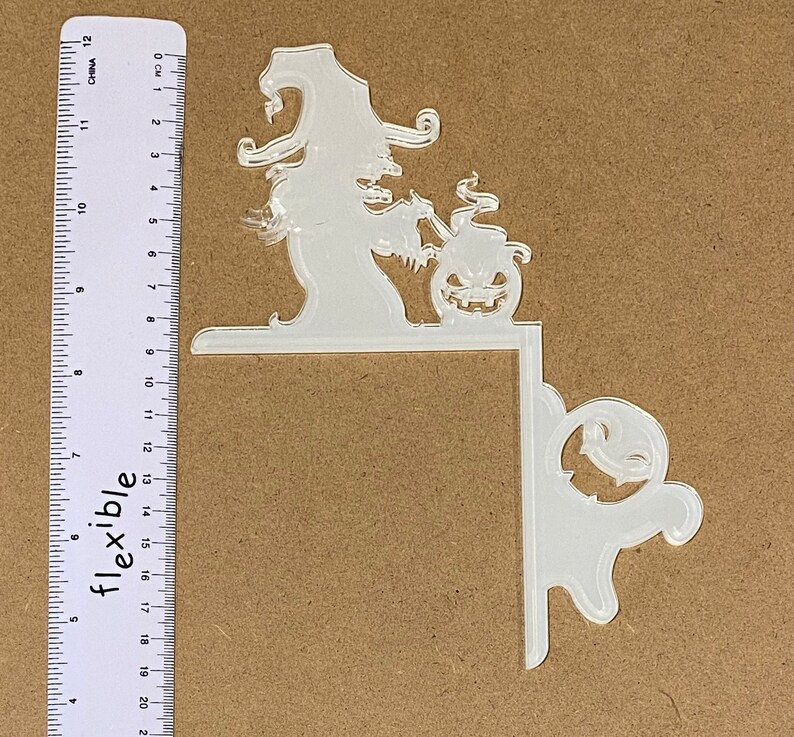 May include: Clear acrylic Halloween corner decoration. The design includes a witch with a pointed hat, a jack-o'-lantern, and a ghost. The decoration is designed to fit around a corner and is approximately 20 cm long.
