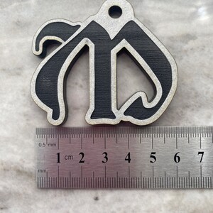 May include: Black wooden ornament shaped like the letter "M" with a white outline. The ornament is approximately 4 cm wide and is measured against a ruler.