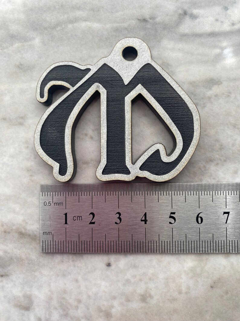 May include: Black wooden ornament shaped like the letter "M" with a white outline. The ornament is approximately 4 cm wide and is measured against a ruler.