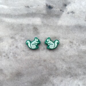 Op de afbeelding: Een paar groene en witte eekhoorn oorbel studs. De eekhoorns kijken elkaar aan en houden noten in hun poten.