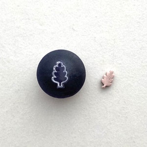 Puede incluir: Un sello redondo negro con un contorno blanco de una hoja de roble. También se muestra una hoja de roble rosa más pequeña.