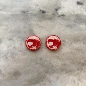Puede incluir: Un par de pendientes redondos rojos con un contorno blanco de una cabeza de león.