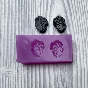 Può includere: Uno stampo in silicone viola con due cavità a forma di cuore. Due pezzi di resina a forma di cuore nero sono mostrati accanto allo stampo.