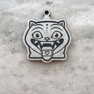 May include: A wooden cat-shaped pendant with a black outline and engraved details. The cat has large eyes, a toothy grin, and a heart-shaped design on its tongue. The pendant has a small hole at the top for hanging.