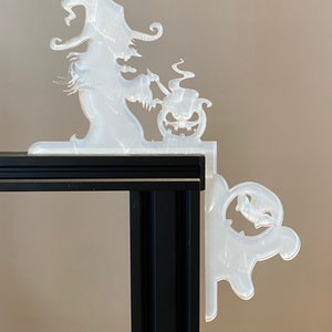 May include: A clear, corner-mounted Halloween decoration featuring a witch stirring a cauldron and a jack-o'-lantern. A ghost figure is on the other side. The decoration is made of a translucent material and is designed to fit over a corner.