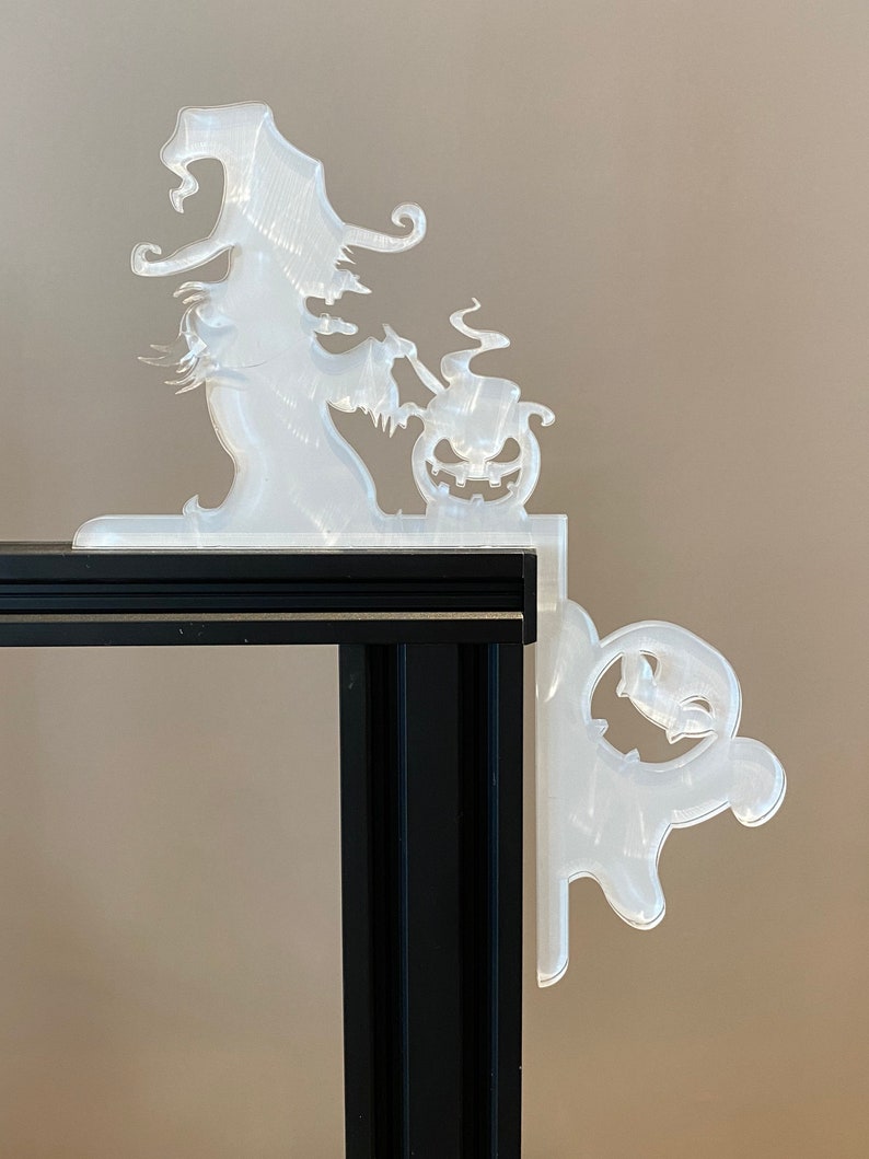 May include: A clear, corner-mounted Halloween decoration featuring a witch stirring a cauldron and a jack-o'-lantern. A ghost figure is on the other side. The decoration is made of a translucent material and is designed to fit over a corner.