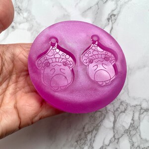 May include: A close-up of a purple silicone mold with two mushroom-shaped impressions. The mold is designed for making earrings.