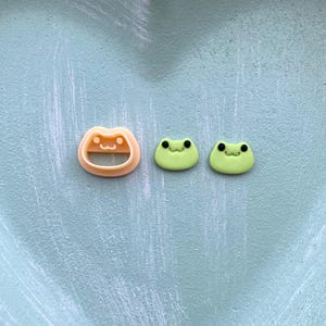 Op de afbeelding: Een lichtoranje kikker-vormige koekjesvorm naast twee groene kikker-vormige decoraties. De koekjesvorm heeft een lachend gezichtsontwerp. De kikkerdecoraties hebben zwarte ogen en lachende monden. De objecten liggen op een lichtblauwe textuur.