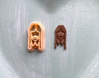 May include: Two cookie cutters, one is a light brown plastic outline of a person with long hair and the other is a brown clay cutout of a person with long hair.