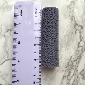 May include: A grey cylindrical roller with a repeating pattern of abstract shapes is shown next to a ruler. The roller is approximately 10 cm long. The ruler is marked with both inches and centimetres.