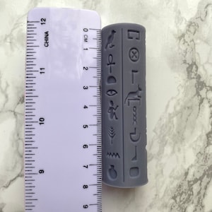 May include: A grey cylindrical object featuring ancient Egyptian hieroglyphs is displayed next to a ruler. The object is approximately 10 cm long. The ruler is marked with both centimetres and inches. The object is a light grey colour.
