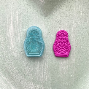 Pode incluir: Um conjunto de duas bonecas russas, uma é um molde de plástico transparente e a outra é uma boneca rosa sólida. Ambas as bonecas têm um design tradicional de boneca russa.