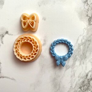 May include: Three cookie cutters, one in the shape of a wreath with a bow, one in the shape of a wreath, and one in the shape of a bow. The wreath cookie cutters are beige and the bow cookie cutter is blue.