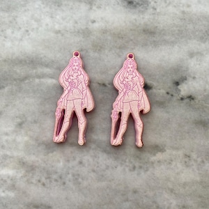 May include: Two pink laser-cut acrylic earrings featuring a stylized female figure with long hair, a cape, and a staff. The earrings are in a standing pose, with a small loop at the top for hanging. The background is a light gray marble.