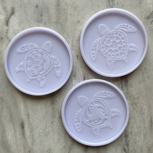 May include: Three light purple, round coasters with raised sea turtle designs. Each coaster features a different turtle design, including floral, heart, and dotted patterns. The coasters are made of a smooth material.