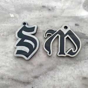 May include: Two black wooden ornaments shaped like the letters "S" and "D" in a gothic font. The letters are painted black with a silver glitter finish.