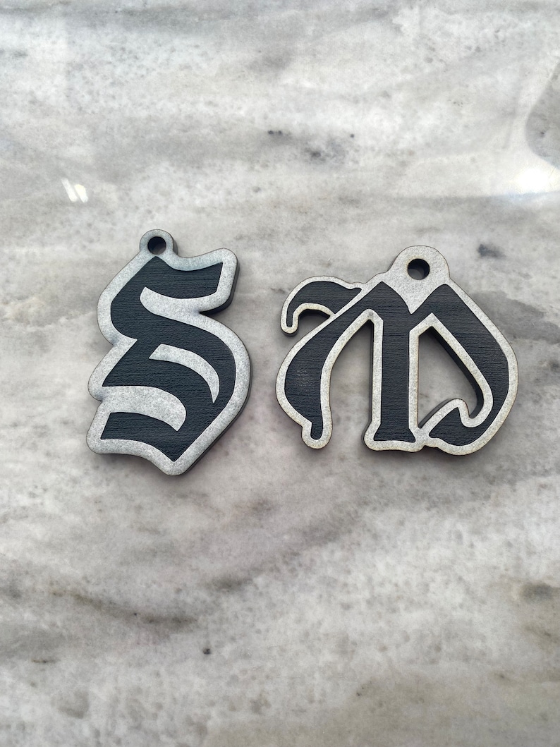 May include: Two black wooden ornaments shaped like the letters "S" and "D" in a gothic font. The letters are painted black with a silver glitter finish.
