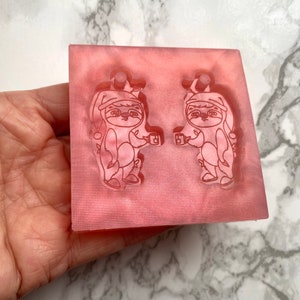 May include: A pink silicone mold with two impressions of a sloth wearing a Santa hat and holding a mug. The sloth is outlined in white.