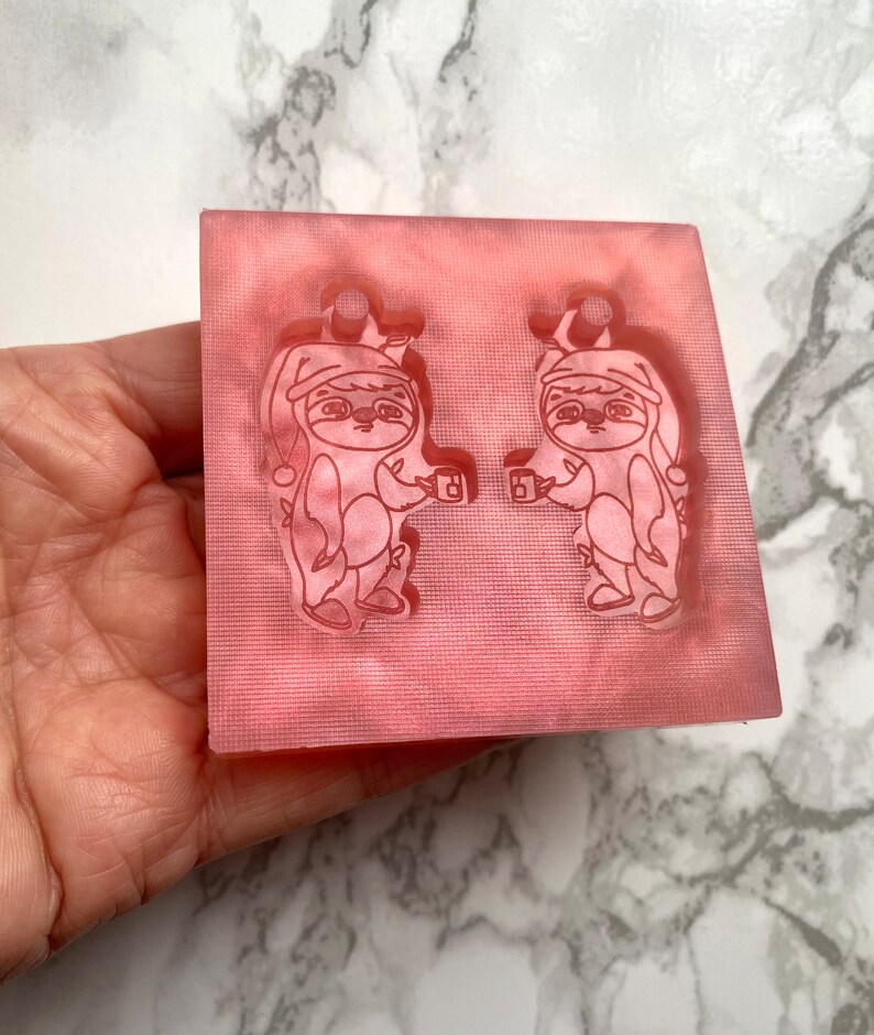 May include: A pink silicone mold with two impressions of a sloth wearing a Santa hat and holding a mug. The sloth is outlined in white.