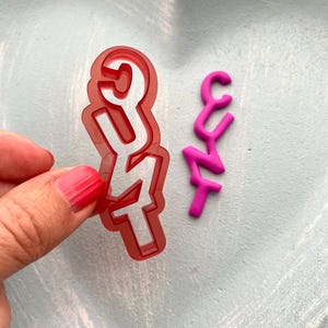 Op de afbeelding: Een rode plastic koekjesvorm in de vorm van het woord "CUT" wordt vastgehouden naast een kleine, roze, driedimensionale versie van het woord. De vorm is op een lichtblauwe ondergrond.