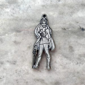 May include: A silver-colored charm featuring a stylized illustration of a female figure with long flowing hair, a cape, and a sword. The charm has a hole at the top for attachment and is likely made of metal.