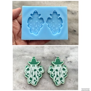 Puede incluir: Dos pendientes de fantasma verde y blanco con un sombrero verde. Los pendientes están hechos de resina y tienen forma de fantasma con un sombrero verde. Los fantasmas tienen ojos blancos y una boca blanca.