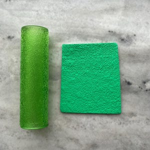 Op de afbeelding: Een getextureerd, cilindrisch, groen glazen object en een vierkant, getextureerd, groen object worden weergegeven op een marmeren oppervlak. Het glazen object is een lange, slanke cilinder. Het vierkante object heeft een ruwe, bobbelige textuur.